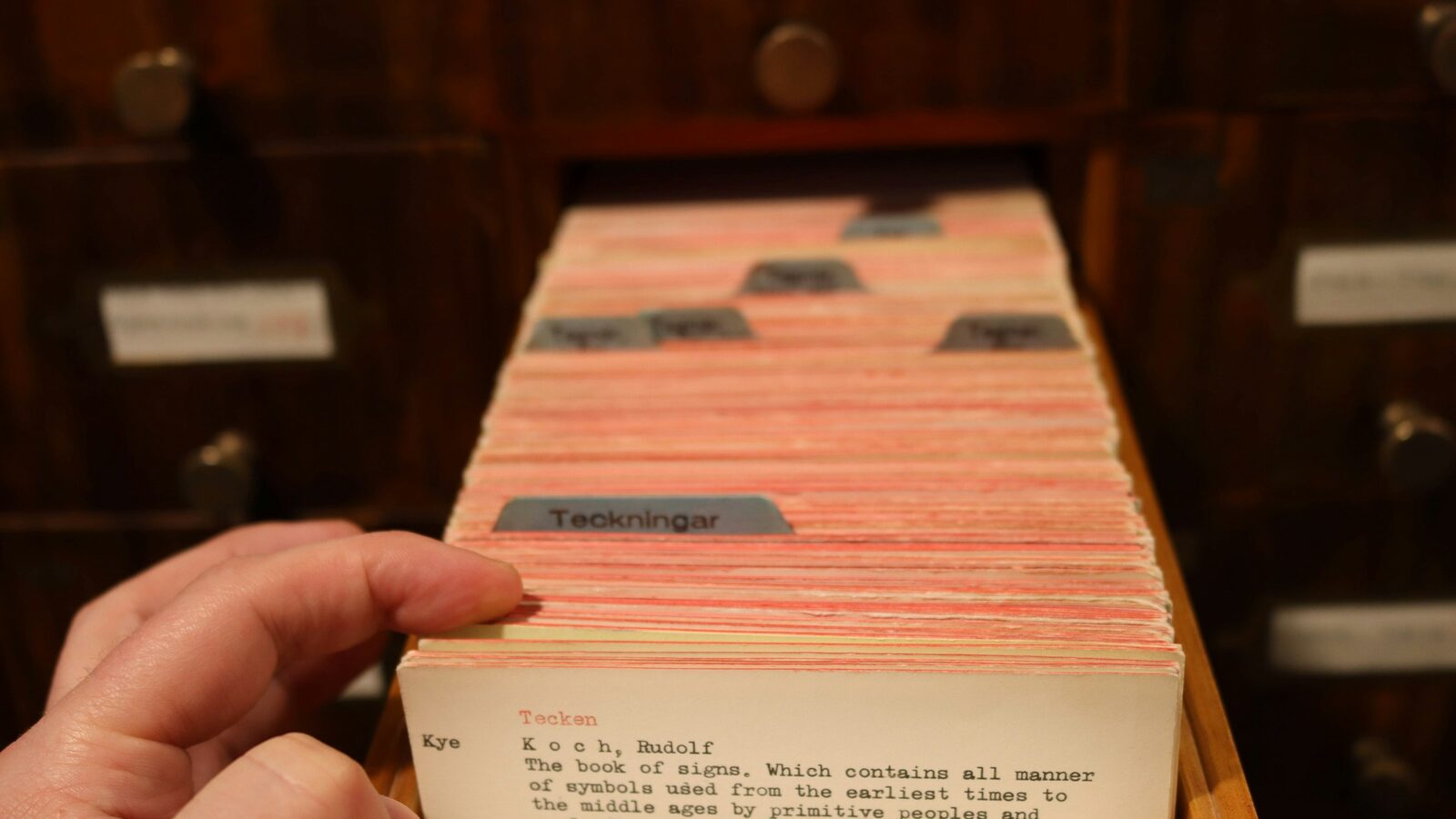 A hand holding a typed index card at the front of an open library card catalog drawer