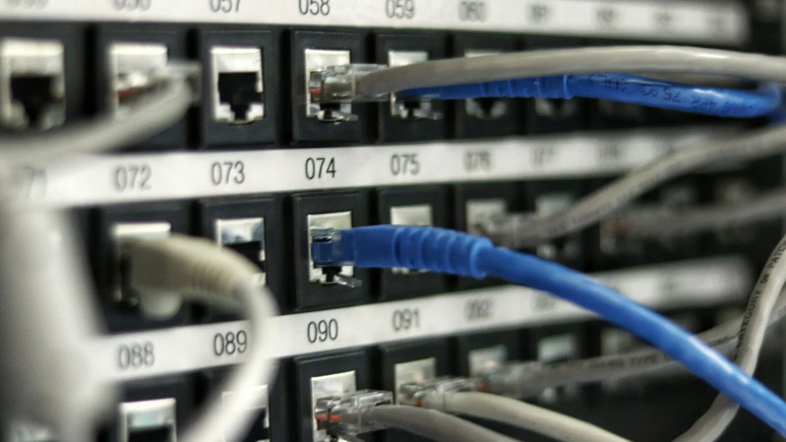 A network patch panel with numbered ports, one blue cable plugged into port 074 among grey ones